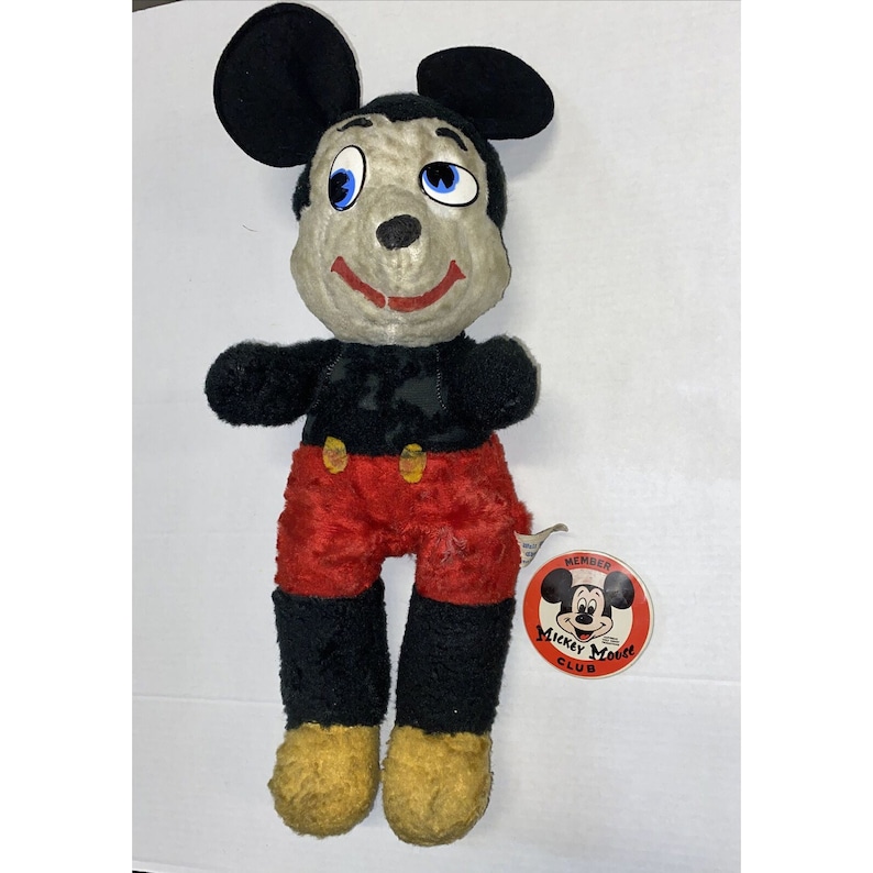 May include: Vintage Mickey Mouse plush toy with black fur, red trousers, and yellow shoes. The toy has a worn and faded appearance. It has a Mickey Mouse Club membership tag attached.