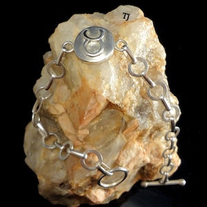 May include: Silver bracelet with a circular charm featuring the Taurus zodiac symbol. The bracelet has a toggle clasp.