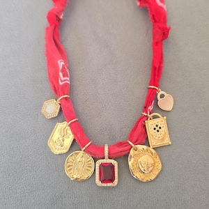 Op de afbeelding: Een ketting van een rode stoffen bandana met een centrale rechthoekige rode edelsteen hanger omringd door goudkleurige bedels, waaronder een zon, een religieuze figuur, een hart en een rechthoekig frame aan de geknoopte stoffen band.