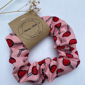 Puede incluir: Un scrunchie rosa con un estampado de piruletas rojas y blancas. El scrunchie tiene una etiqueta marrón que dice "Handmade for you" y "Going Wild" con un corazón alrededor del texto. La etiqueta también dice "Thanks a lot!"