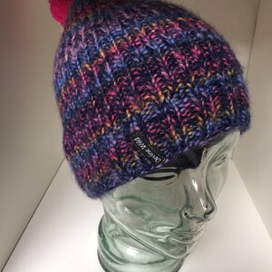 Könnte beinhalten: Eine handgestrickte Mütze mit einem bunten Farbmix aus Blau, Lila, Pink und Orange. Die Mütze hat einen auffälligen pinkfarbenen Bommel und ein kleines schwarzes Etikett mit der Aufschrift "Devine Wild". Die Mütze wird auf einem transparenten Glaskopf präsentiert.
