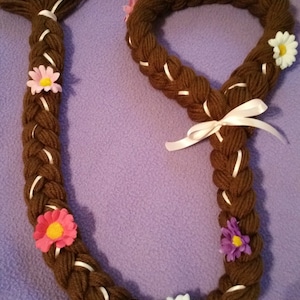 May include: A brown yarn braid with white ribbon accents and colorful felt flowers. The braid is long and has a loop at the top.