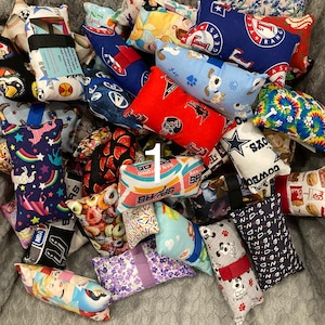 May include: A collection of small, rectangular fabric items with various patterns and designs. Some feature sports team logos, cartoon characters, and other colorful prints. Each item has a black or colored strap.