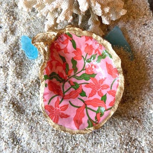 May include: A decorative oyster shell dish with a pink floral pattern and a gold-painted rim. The shell is set on a bed of sand with a piece of coral and blue sea glass.
