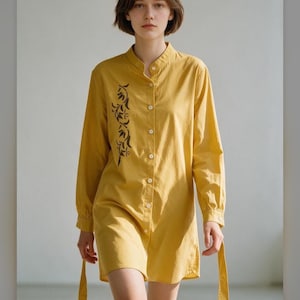 Puede incluir: Un vestido camisero de color amarillo mostaza con mangas largas, cuello mao, tapeta de botones frontal y un patrón de hojas bordado en marrón oscuro en el lado derecho. La prenda incluye mangas largas con puños abotonados y lazos laterales.