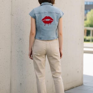 May include: Pale blue denim jacket featuring a red lip design on the back. The jacket has short sleeves and is worn with beige trousers and white trainers. The lip design has a dripping effect.