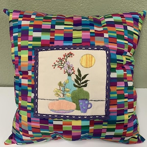 May include: A colorful pillow with a square embroidered design featuring a still life of flowers, vases, and a cup. The pillow has a multi-colored checkered pattern.