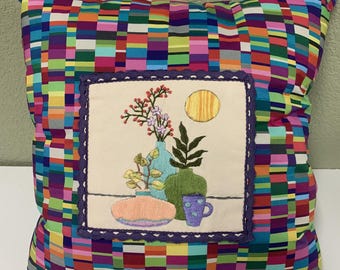Peut inclure: Un oreiller coloré avec un motif brodé carré représentant une nature morte de fleurs, de vases et d'une tasse. L'oreiller a un motif à carreaux multicolores.