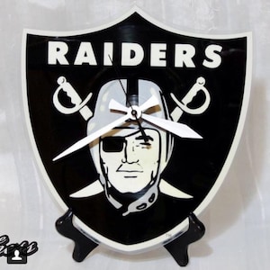 May include: A black and white vinyl record clock featuring the Las Vegas Raiders logo. The clock is shaped like a shield with the Raiders logo in the center. The clock has white hands and a black stand.