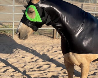 May include: A horse wearing a black, shiny hood with green eye accents and spiky black trim. The horse has a tan coat and is standing in a sandy area with a fence in the background.