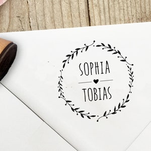 Könnte beinhalten: Ein weißer Umschlag mit einem schwarzen Stempel, der einen Kranz aus Blättern mit den Namen "Sophia" und "Tobias" im Inneren zeigt, die durch ein Herzsymbol getrennt sind.