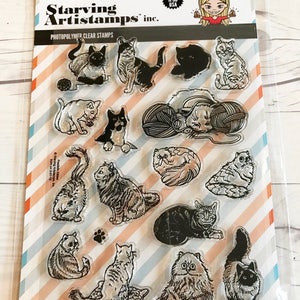 May include: Clear photopolymer stamps featuring various cat illustrations. The stamp set includes images of cats in different poses, with yarn balls, and paw prints. The packaging includes the text "Starving Artistamps inc." and "Made in the USA."