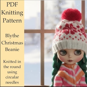 Op de afbeelding: Een pop draagt een wit en rode gebreide kerstmuts met een rode pompon. De muts heeft een sneeuwvlokpatroon. De pop heeft groene vlechten en draagt een roze en oranje gestreepte top. Tekst zegt "PDF Knitting Pattern Blythe Christmas Beanie".