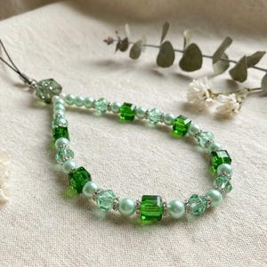 May include: A beaded phone charm featuring a mix of green beads, including cube-shaped and round beads, and clear crystal accents. The charm is strung on a black cord and has a green, faceted bead at the top. The beads are in shades of green and light green.