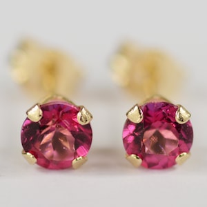 May include: A pair of stud earrings featuring round, faceted, pink gemstones set in gold-colored metal. The earrings have a classic, minimalist design, with the gemstones held in place by four prongs.