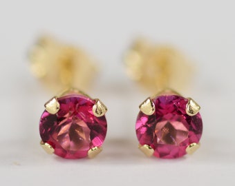 May include: A pair of stud earrings featuring round, faceted, pink gemstones set in gold-colored metal. The earrings have a classic, minimalist design, with the gemstones held in place by four prongs.