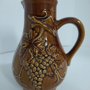 May include: Brown ceramic pitcher with a handle and a cork stopper. The pitcher features a raised relief design of grape vines and leaves. The pitcher has a capacity marking of 0.5L.