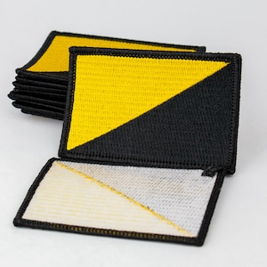 Könnte beinhalten: Ein Stapel schwarzer und gelber Stoff-Patches mit einer diagonalen Linie, die die Farben trennt. Die Patches sind mit schwarzem Garn genäht. Die Patches sind in der Mitte gefaltet und zeigen die weiße Rückseite.