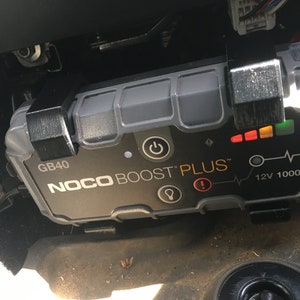 May include: A grey NOCO BOOST PLUS GB40 jump starter with a black power button, a red warning light, and a green, yellow, and red indicator light. The jump starter is labelled "12V 1000A".