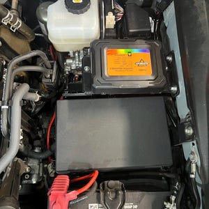 Puede incluir: Un compartimento del motor con varios componentes, incluyendo una batería, una caja de fusibles y un depósito de líquido. La batería tiene una cubierta de terminal roja. Una caja negra con el texto "SWITCH ASSEMBLY" es visible.