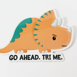 K&ouml;nnte beinhalten: Ein Aufkleber mit einem orangefarbenen und gr&uuml;nen Triceratops-Cartoon mit dem Text "GO AHEAD. TRI ME."