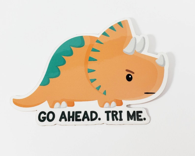 K&ouml;nnte beinhalten: Ein Aufkleber mit einem orangefarbenen und gr&uuml;nen Triceratops-Cartoon mit dem Text "GO AHEAD. TRI ME."
