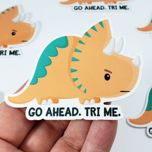 K&ouml;nnte beinhalten: Ein Aufkleber mit einem orangefarbenen und gr&uuml;nen Triceratops-Cartoon mit dem Text "Go Ahead. Tri Me."