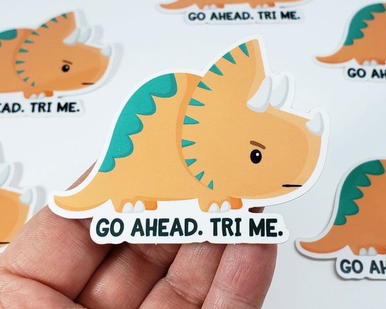 K&ouml;nnte beinhalten: Ein Aufkleber mit einem orangefarbenen und gr&uuml;nen Triceratops-Cartoon mit dem Text "Go Ahead. Tri Me."