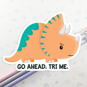 K&ouml;nnte beinhalten: Ein Aufkleber mit einem orangefarbenen und t&uuml;rkisfarbenen Triceratops-Cartoon mit dem Text "GO AHEAD. TRI ME."