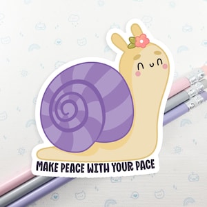 May include: A die-cut sticker featuring a cartoon snail with a purple shell and a flower on its head. The sticker has the text "MAKE PEACE WITH YOUR PACE" in black. Pencils in pink, gray, and purple are in the background.