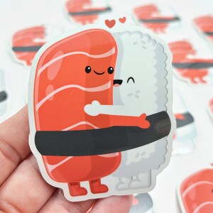 Puede incluir: Una pegatina de dos rollos de sushi abraz&aacute;ndose. El rollo de salm&oacute;n es rojo con una envoltura de alga negra y el rollo de arroz blanco tiene una envoltura de alga negra. Ambos rollos tienen caras lindas y sostienen corazones.