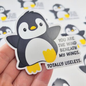 以下が含まれることがあります： 「You are the wind beneath my wings. Totally useless.」というテキストが書かれた、漫画のペンギンが描かれたステッカーです