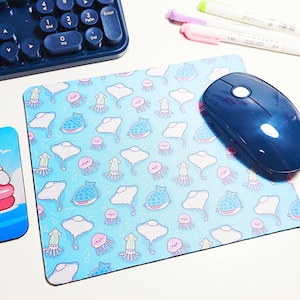 May include: A blue computer mouse rests on a matching mousepad with a repeating pattern of cartoon sea creatures, including stingrays, squid, and jellyfish. A blue keyboard and a coaster with a cartoon character are also visible.