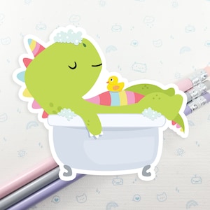 May include: A green dinosaur with a rainbow tail is taking a bath in a white bathtub. The dinosaur has a yellow rubber ducky in the tub with it. The dinosaur is smiling and has its eyes closed.