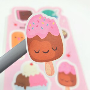 May include: A sticker featuring a cartoon ice cream bar with a pink top, chocolate base, and colorful sprinkles. The ice cream has a smiling face and is on a wooden stick. Other ice cream stickers are in the background.