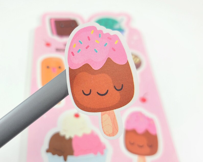 May include: A sticker featuring a cartoon ice cream bar with a pink top, chocolate base, and colorful sprinkles. The ice cream has a smiling face and is on a wooden stick. Other ice cream stickers are in the background.