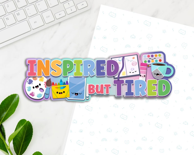 May include: A colorful graphic design with the text "Inspired But Tired" and cartoon images of art supplies, including a paint palette, crayons, a sketchbook, and a coffee cup.