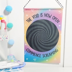 May include: A rectangular wall hanging with a rainbow gradient background and a black spiral design. The text "THE VOID IS NOW OPEN" and "COMMENCE SCREAMING" are printed on the fabric. The hanging is suspended by a black and white cord.