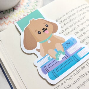 May include: A brown and white cartoon dog bookmark with a blue collar and a green ball in its paw. The dog is sitting on top of a stack of colorful books.
