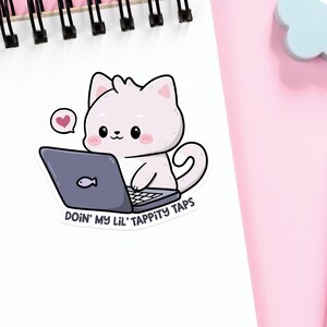 K&ouml;nnte beinhalten: Eine wei&szlig;e Katze mit rosa Wangen sitzt an einem Laptop. Die Katze tr&auml;gt ein rosa Herz &uuml;ber dem Kopf. Der Laptop hat einen blauen Bildschirm mit einem Fisksymbol. Der Text unter der Katze lautet "Doin' my lil' tappity taps".