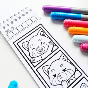 May include: A spiral-bound coloring book with red panda illustrations and the text "COLOR TEST." Several colorful markers in purple, blue, pink, orange, white, and teal are arranged next to the book, ready for use.