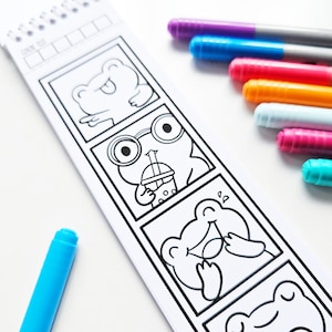 May include: A white coloring book featuring cartoon frog illustrations, with a spiral binding and a selection of colorful markers. The frog drawings depict various expressions. A blue marker is in the foreground.