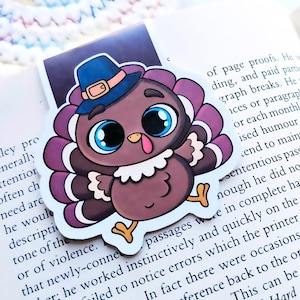May include: A whimsical turkey bookmark with a blue pilgrim hat and large blue eyes. The cartoon turkey has a brown body, purple and white feathers, and yellow feet. The bookmark is placed on a book page.