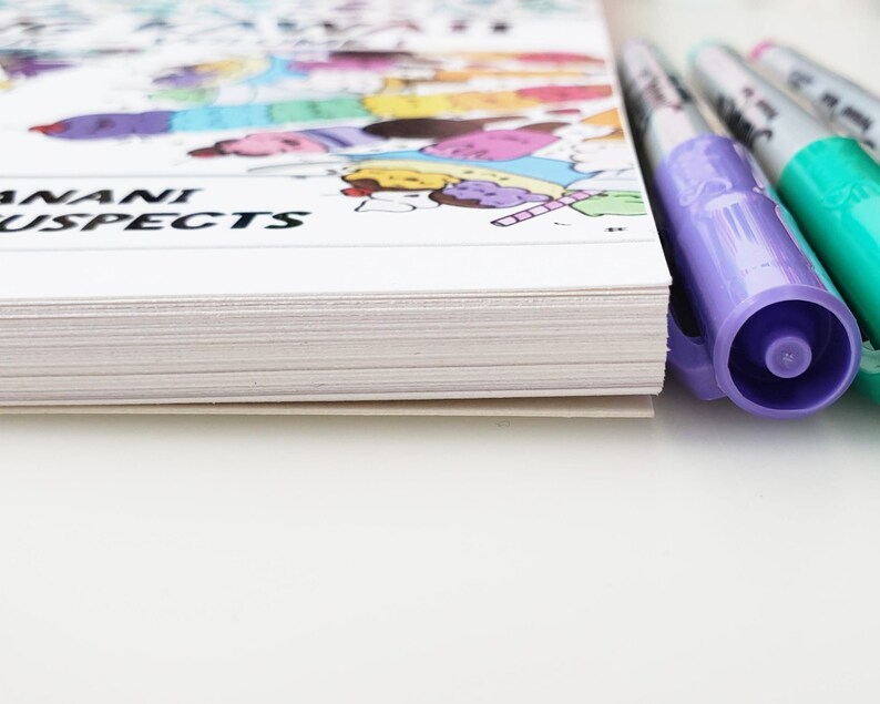 May include: A white book with the text "Anani Suspects" on the cover. The book is closed and lying on a white surface. There are three colorful markers next to the book.