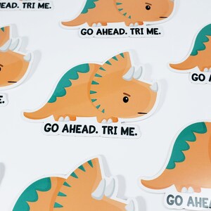 K&ouml;nnte beinhalten: Ein Set mit sechs bunten Aufklebern, die jeweils ein Comic-Triceratops mit dem Text "Go Ahead. Tri Me." zeigen.