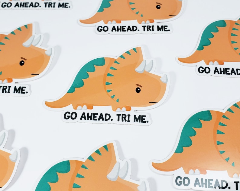 K&ouml;nnte beinhalten: Ein Set mit sechs bunten Aufklebern, die jeweils ein Comic-Triceratops mit dem Text "Go Ahead. Tri Me." zeigen.
