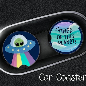 May include: Two car coasters with a colorful design. One coaster features a cartoon alien in a spaceship with a rainbow beam. The other coaster has a blue and green design with the text "Tired of this planet".