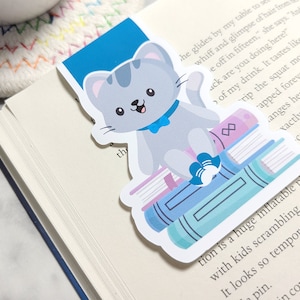 May include: A gray cat with a blue bow tie sits on a stack of colorful books. The cat is smiling and has a white paw sticking out. The bookmark is on a white book with text.