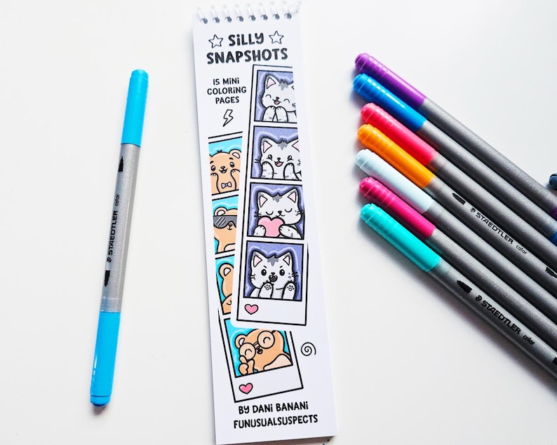 May include: A collection of colorful art supplies, including a blue and silver Staedtler dual-tip marker and several colored Staedtler pens. A small spiral-bound coloring book titled "Silly Snapshots" features cartoon animal illustrations.