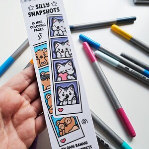May include: A hand holds a bookmark with the text "Silly Snapshots" and "15 Mini Coloring Pages." The bookmark features cartoon cats and bears in polaroid-style frames. Several gray and silver colored markers with different colored tips are scattered around the bookmark.